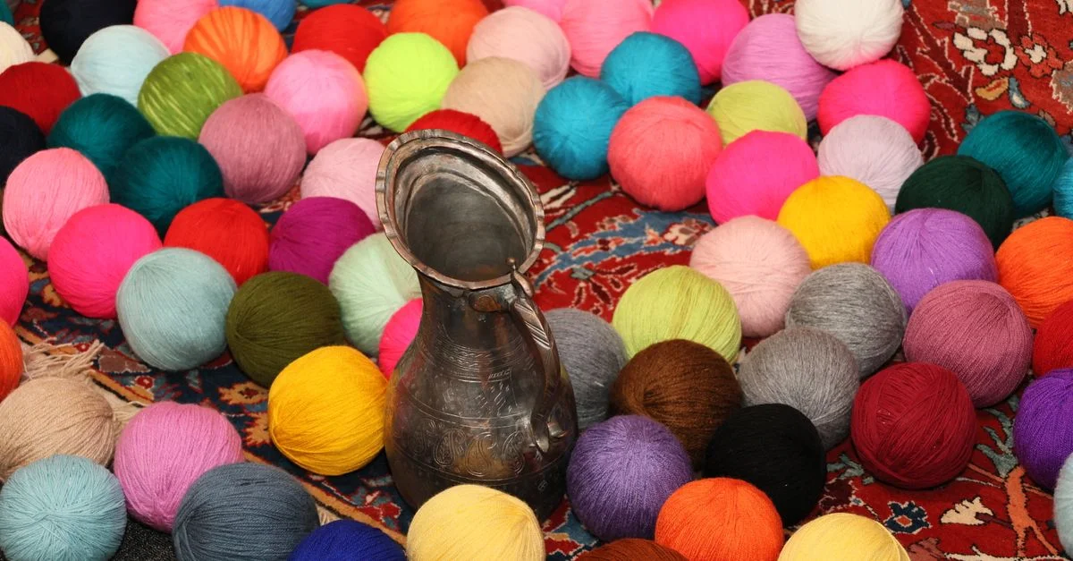 Des pelotes de laine colorées et une cruche en métal sur un tapis à motifs traditionnels, créant une scène vivante et artistique.